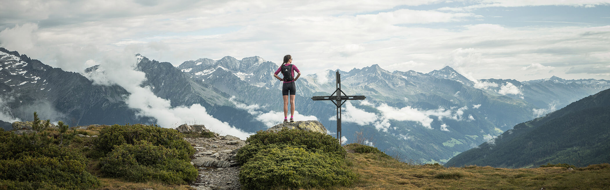 Young woman stands at the summit cross, behind her a monstrous mountain panorama - OLM Nature Escape
