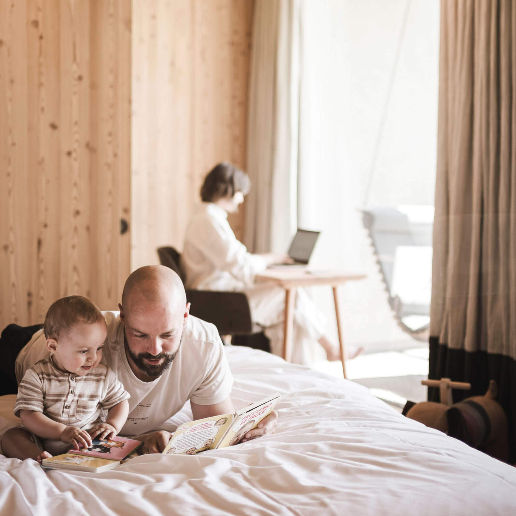Padre e figlio che leggono un libro, la mamma che lavora al portatile sullo sfondo